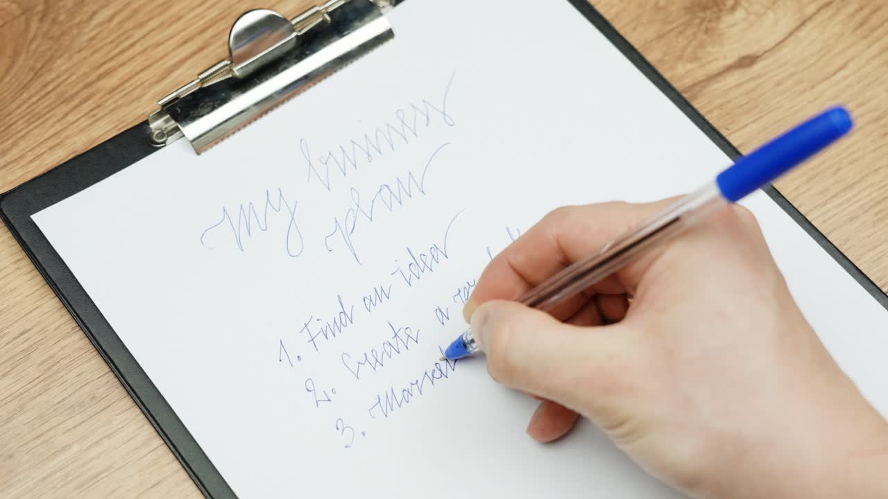 Close-up of a hand writing a business plan on a clipboard using a blue pen with phrases like "Find an idea" and "Search market" are visible on the list, suggesting a creative planning session.