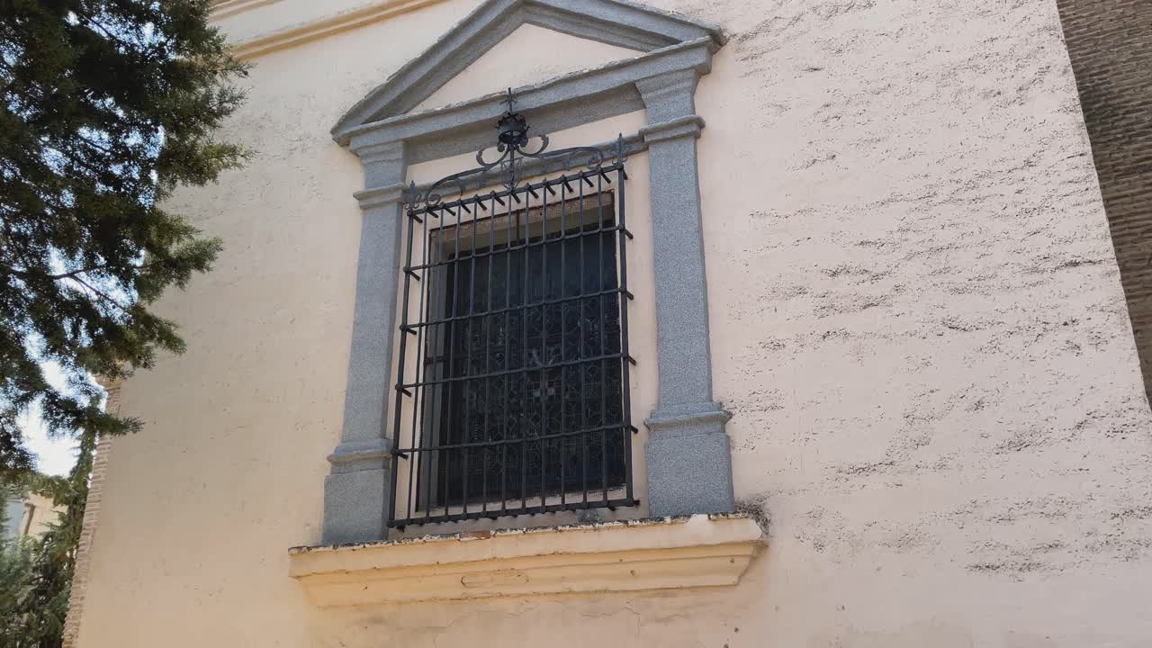 Inquadratura della facciata posteriore di una piccola cappella. Un'ampia finestra con antiche grate in ferro battuto è incorniciata da colonne decorative. Mentre la telecamera si solleva, appaiono il tetto triangolare e un oculo rotondo.