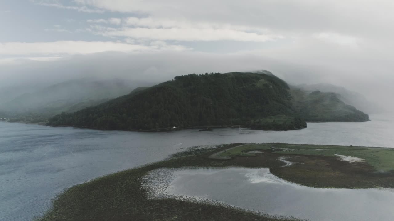 Wide drone shot of a cloud covered coastal mountain with the mist and cloud rolling over the sea and mountain peak in Scotland