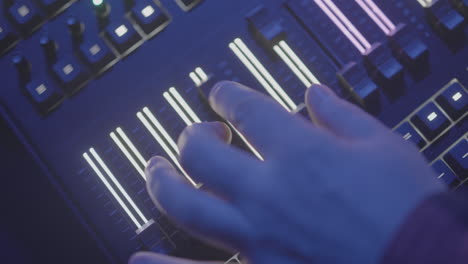 male hand above an illuminated audio or light mixing panel that is sliding up and down a lit slider in slowmotion in a dark event area log