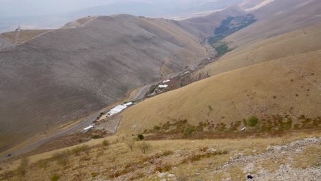 Mirando-Hacia-Abajo-Desde-La-Cima-De-Una-Montaña-A-Una-Carretera-Solitaria-En-El-Valle-De-Abajo-Con-Un-Coche-Conduciendo-Por-La-Carretera