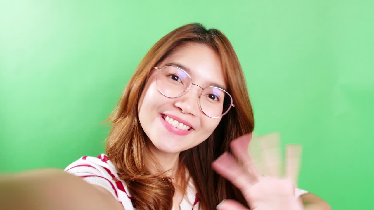 Portrait of a selfie Asian woman with eyeglasses is smiling with laughter and waving her hand being positive emotions on video call online on chroma key green screen