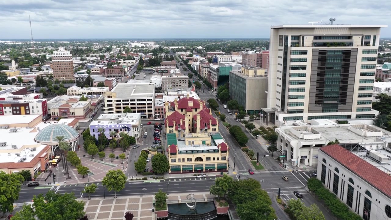 Menara beratap merah yang berornamen menjadi pusat plaza pusat kota Stockton, California, AS, menghadap ke jalur air, gedung perkantoran bertingkat modern, jalan-jalan yang dipenuhi pepohonan, lalu lintas ringan di bawah. Pengambilan gambar drone udara yang naik