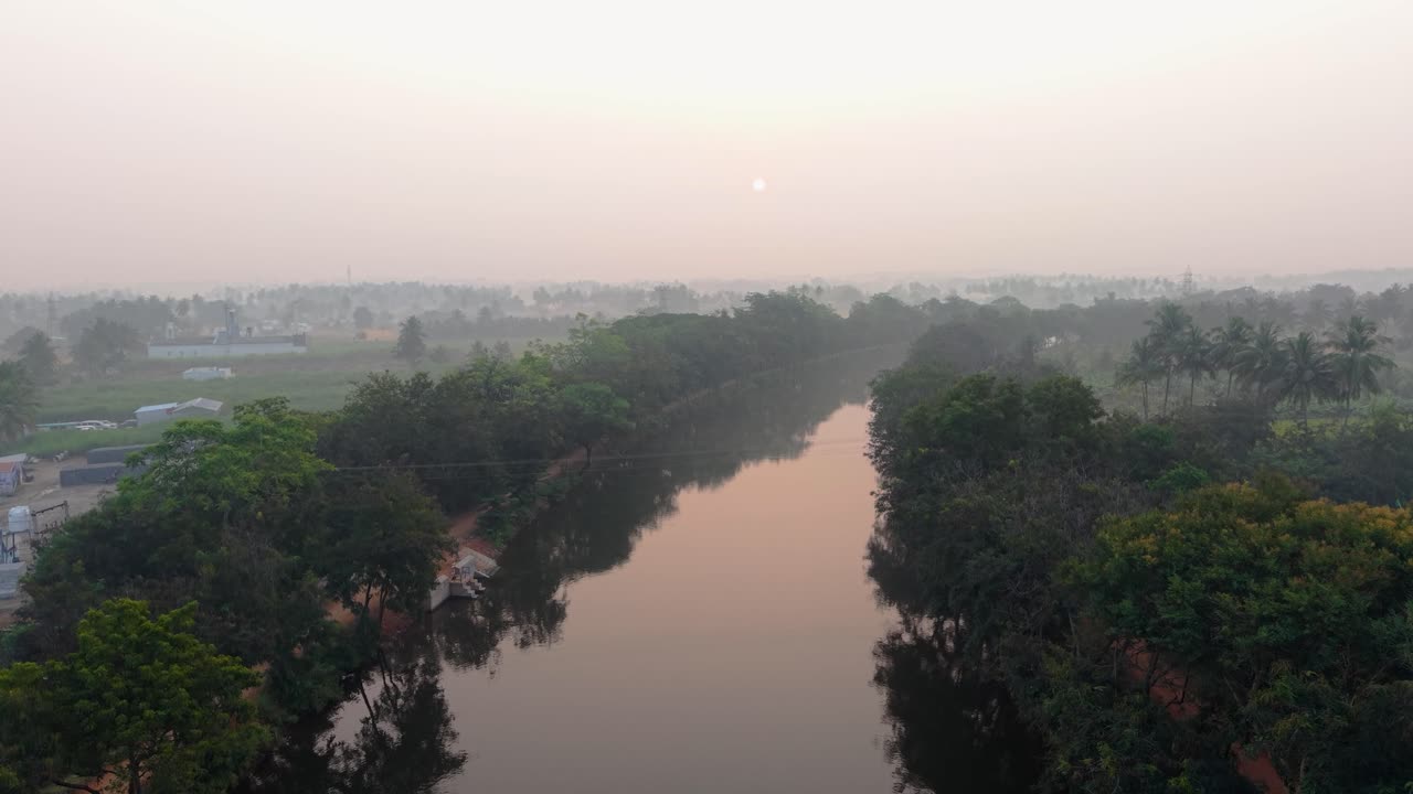 Tranquillo canale che scorre tra rigogliosi alberi verdi all'alba, riflettendo la tenue luce del mattino sull'acqua calma. Sereno paesaggio rurale con simmetria naturale, atmosfera nebbiosa e ambiente di campagna.
