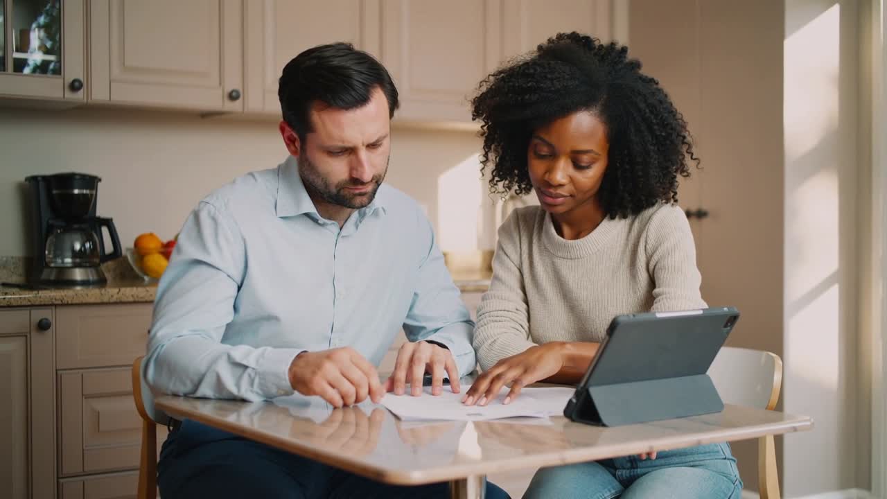 Spreading paperwork, couple at kitchen nook pointing, turning pages and checking tablet for review