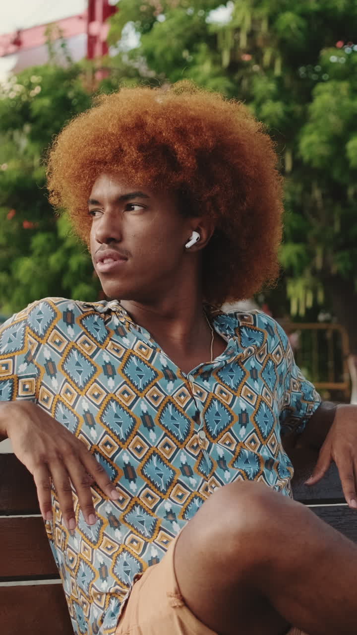 Giovane uomo con capelli afro che si rilassa su una panchina del parco ascoltando musica