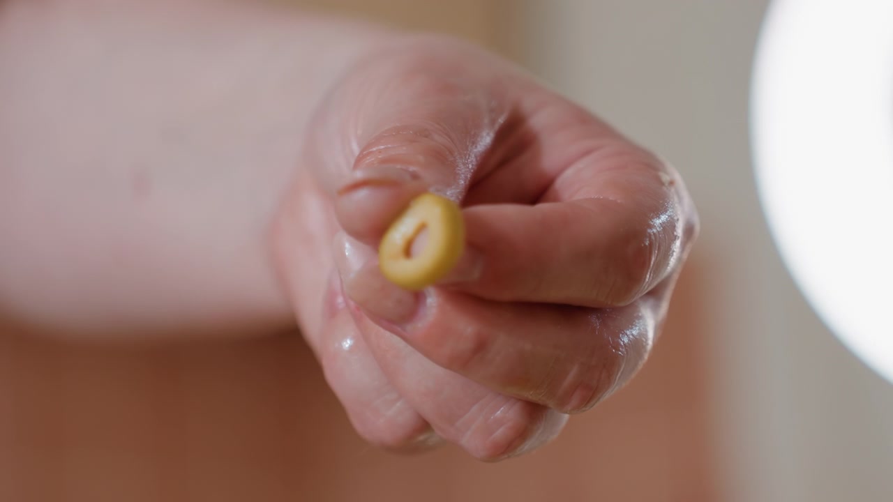 Close view of wet hand shaping tortellini, pinching soft pasta between thumb and forefinger under warm indoor light, showing careful cooking preparation on kitchen surface with glossy dough and
