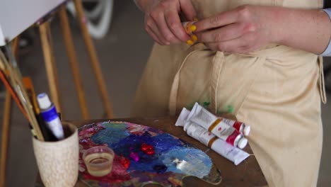 Close-up-footage-caucasian-female-artist-in-apron-squeezing-beige-paint-from-tube-and-putting-on-palette.-Woman,-creative-painter-getting-ready-to-work-over-painting.-Empty-canvas