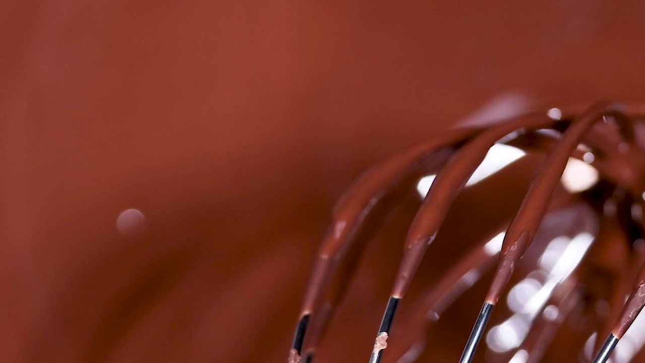 A detailed view of a whisk coated with glossy melted chocolate, highlighting its smooth texture and shine.