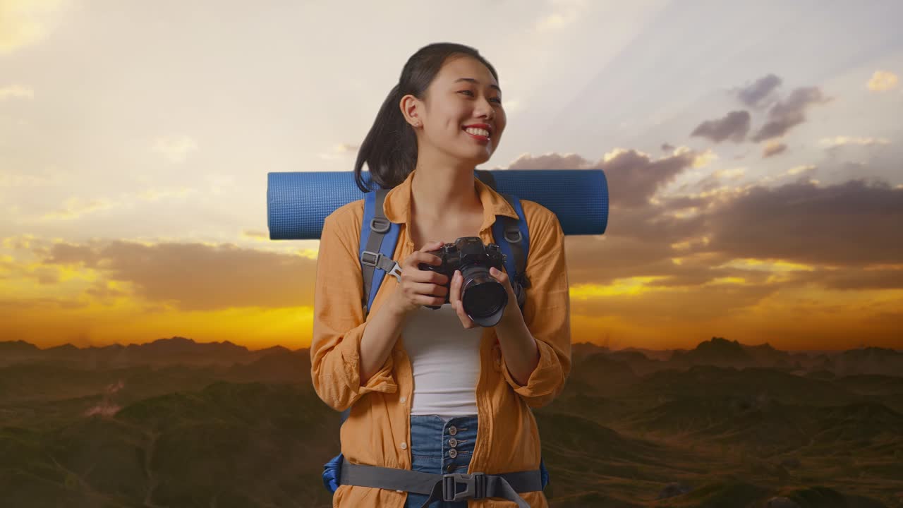 excursionista asiática con mochila de montañismo sonriendo y sosteniendo una cámara en sus manos luego mirando a su alrededor mientras está de pie en la cima de la montaña durante la hora del atardecer