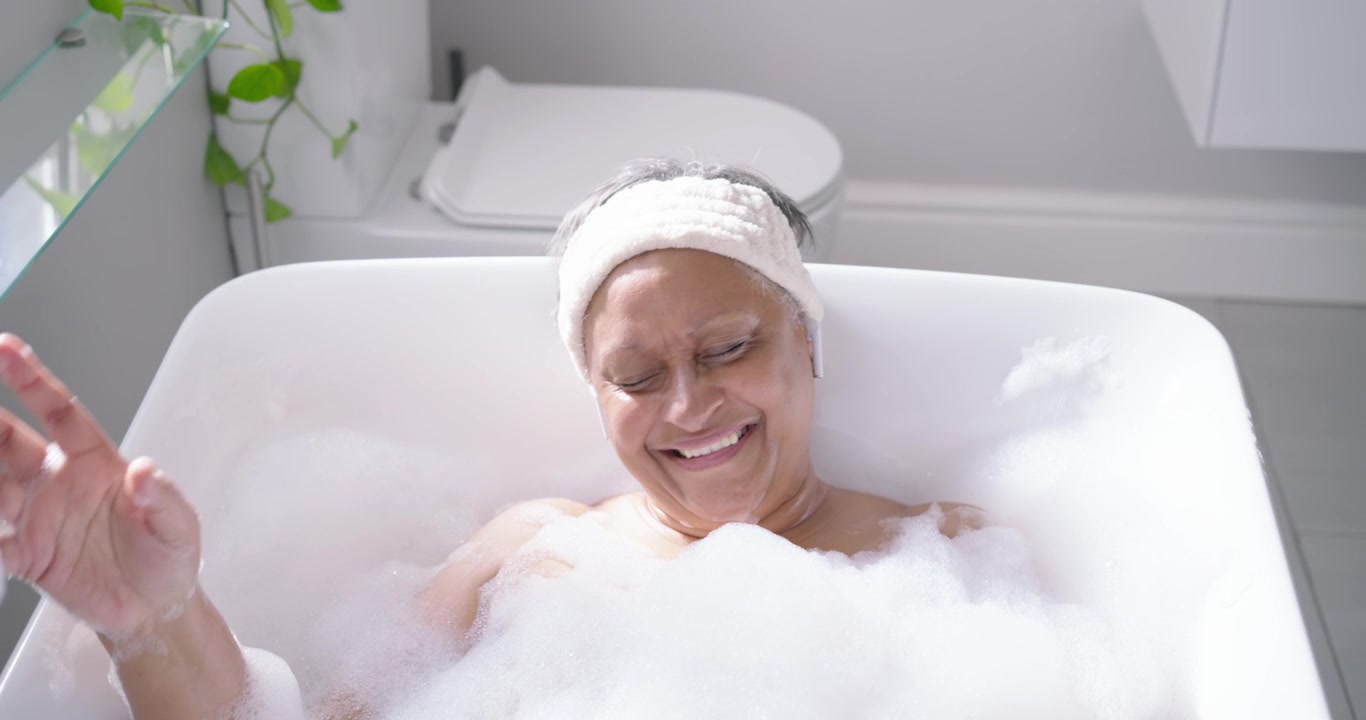 Donna anziana afroamericana che si immerge nella vasca da bagno con una fascia in schiuma per la calma, sorridendo e gesticolando.