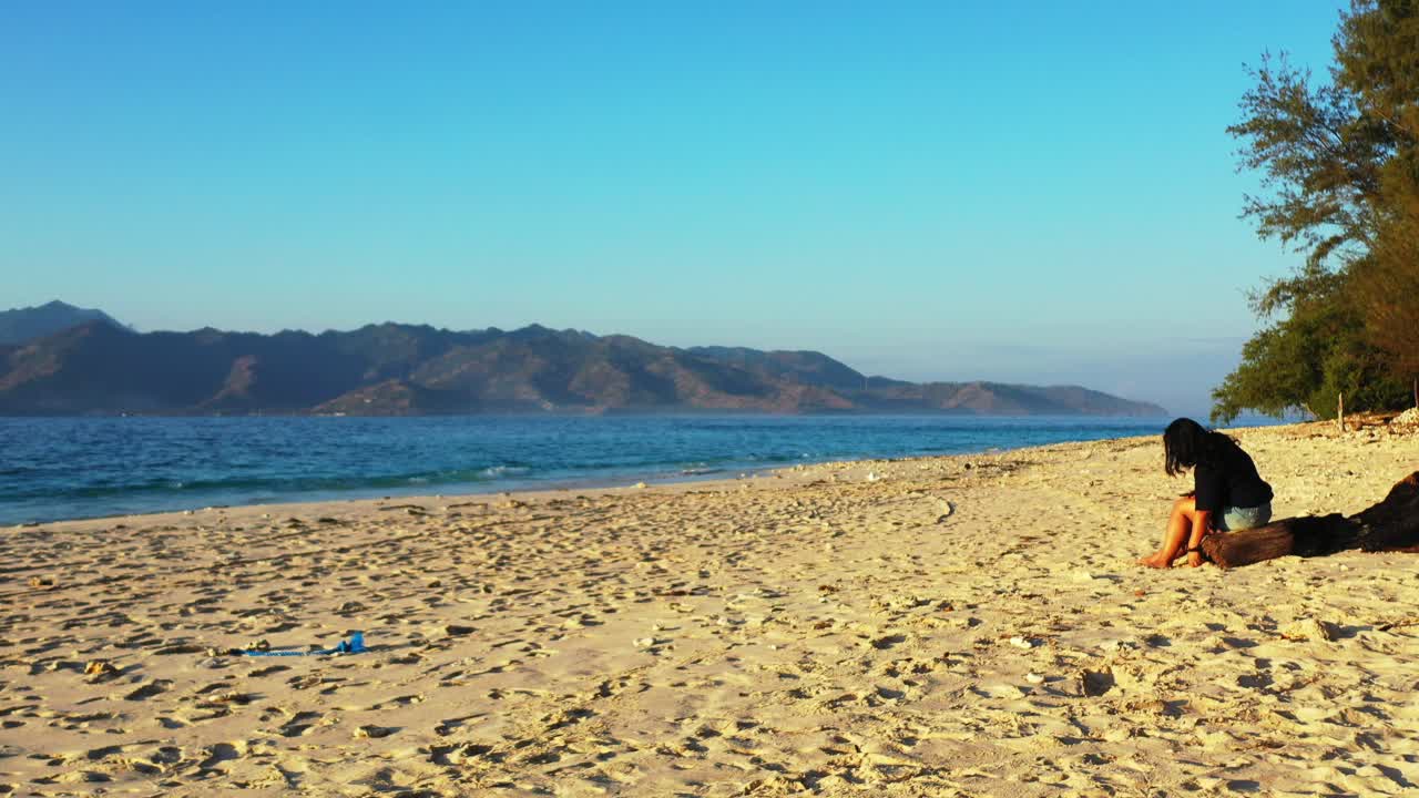 Young woman sitting on dried trunk playing with white sand of exotic beach, watching beautiful blue seascape under bright sky, Bali