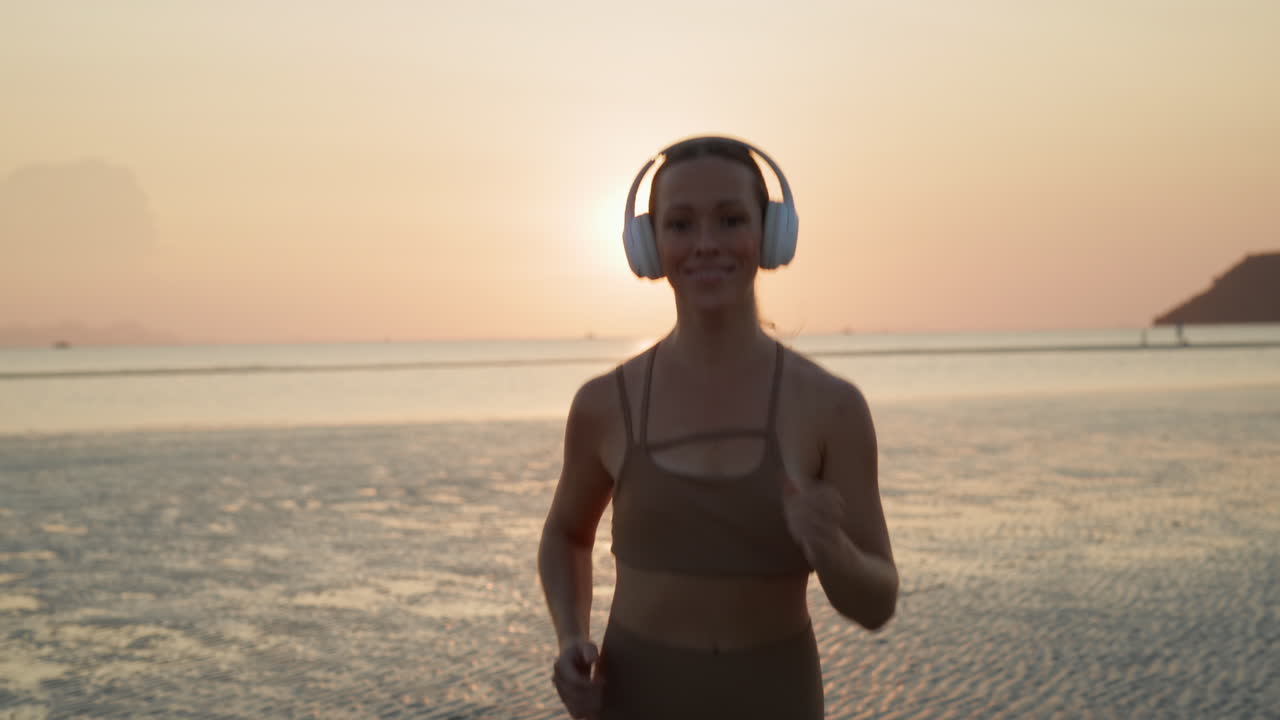 Donna felice che corre sulla spiaggia con le cuffie al tramonto