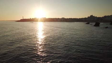 Drone-flying-over-shimmering-waters-with-Biarritz-coast-and-lighthouse-in-background-at-sunset,-France