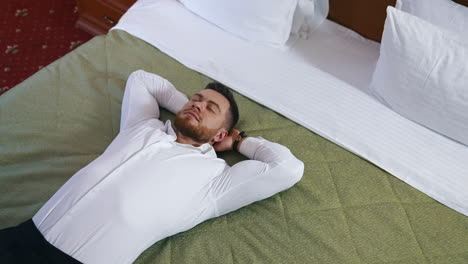 young businessman relaxing on bed. handsome man in white shirt is lying on bed enjoying rest alone in a hotel room. vacation during business trip.