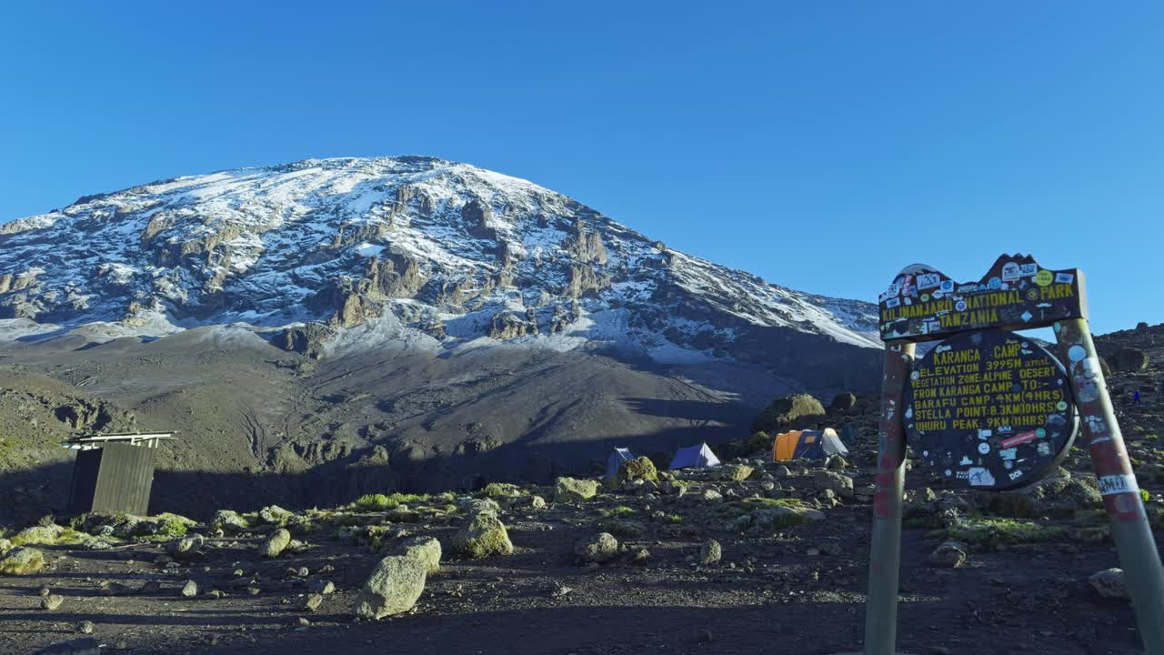 Panning shot captures snow covered Mount Kilimanjaro under clear blue sky, with a Karanga Camp trail sign and rocky terrain at base