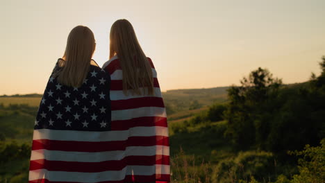 a mother with a teenage daughter with an american flag on their shoulders look at the sunset over a picturesque valley