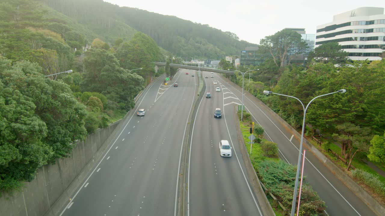 A wide shot high above the traffic driving on the highway leading to and from Wellington city, New Zealand. Someone in a white Tesla realizes they are in the wrong lane and makes a late correction.