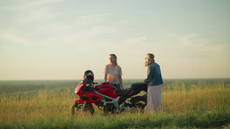 two sisters are cleaning a red motorcycle in an open grassy field, one focuses on wiping the tank, while the other cleans the seat
