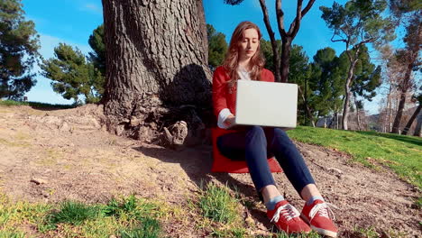 una mujer feliz, atractiva, joven y caucásica en el parque, escribiendo en su computadora portátil debajo de un árbol
