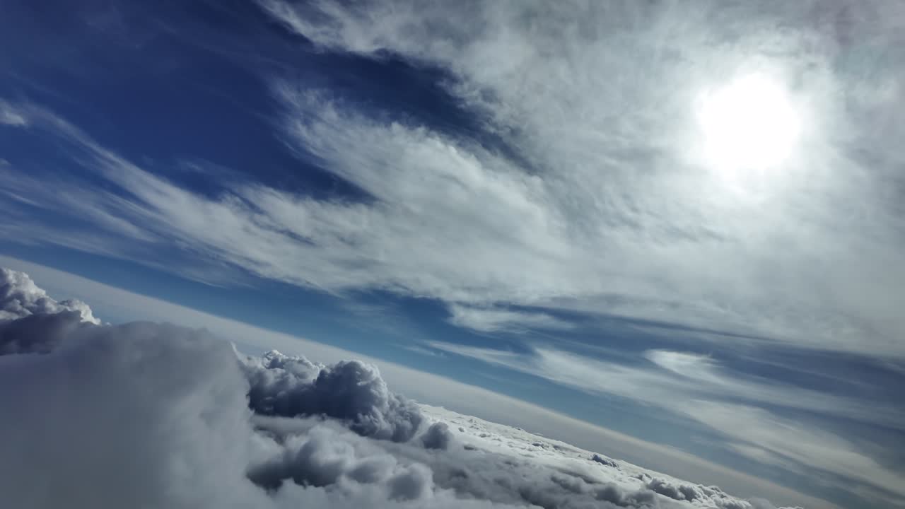 an immervive aerial cloudscape view taken from a jet cockpit flying above cottony clouds in a left turn under a shining sun veiled by ethereal cirrus clouds.