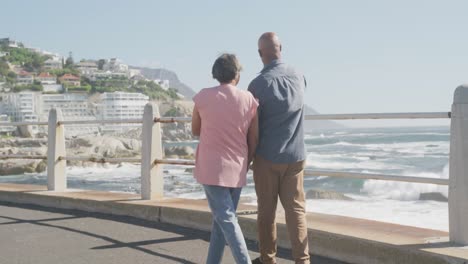 Happy-senior-african-american-couple-walking-along-promenade-by-the-sea,-slow-motion