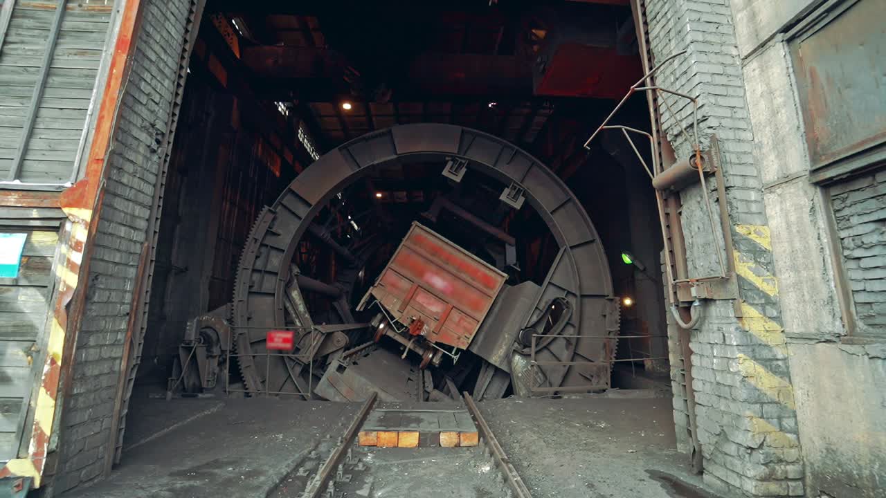 Wagon tipper is spinning with a wagon of coal inside it and pouring out fossil in th building of plant. Camera motion to forward. Close-up.