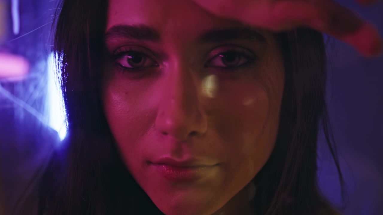 Primo piano volto femminile, luci neon nella città notturna. Donna che si ripara gli occhi da un'insegna luminosa.