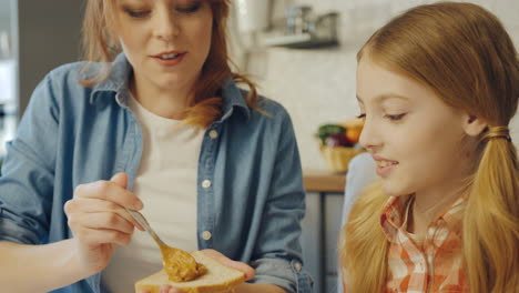 Young-attractive-mother-spreading-a-peanut-butter-on-the-bread-and-talking-with-her-teen-daughter-while-they-sitting-at-the-table-at-the-kitchen.-Close-up.-Inside