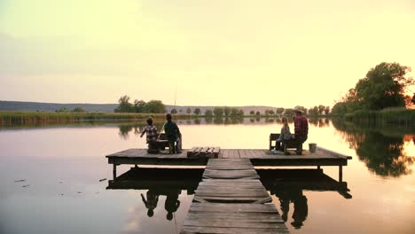 distant rear view teen boys and girl sitting with their grandfather on the lake pier and fishing together