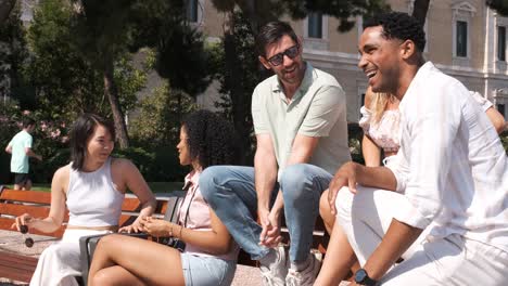 relaxed happy multiethnic friends talking while sitting on bench in city park