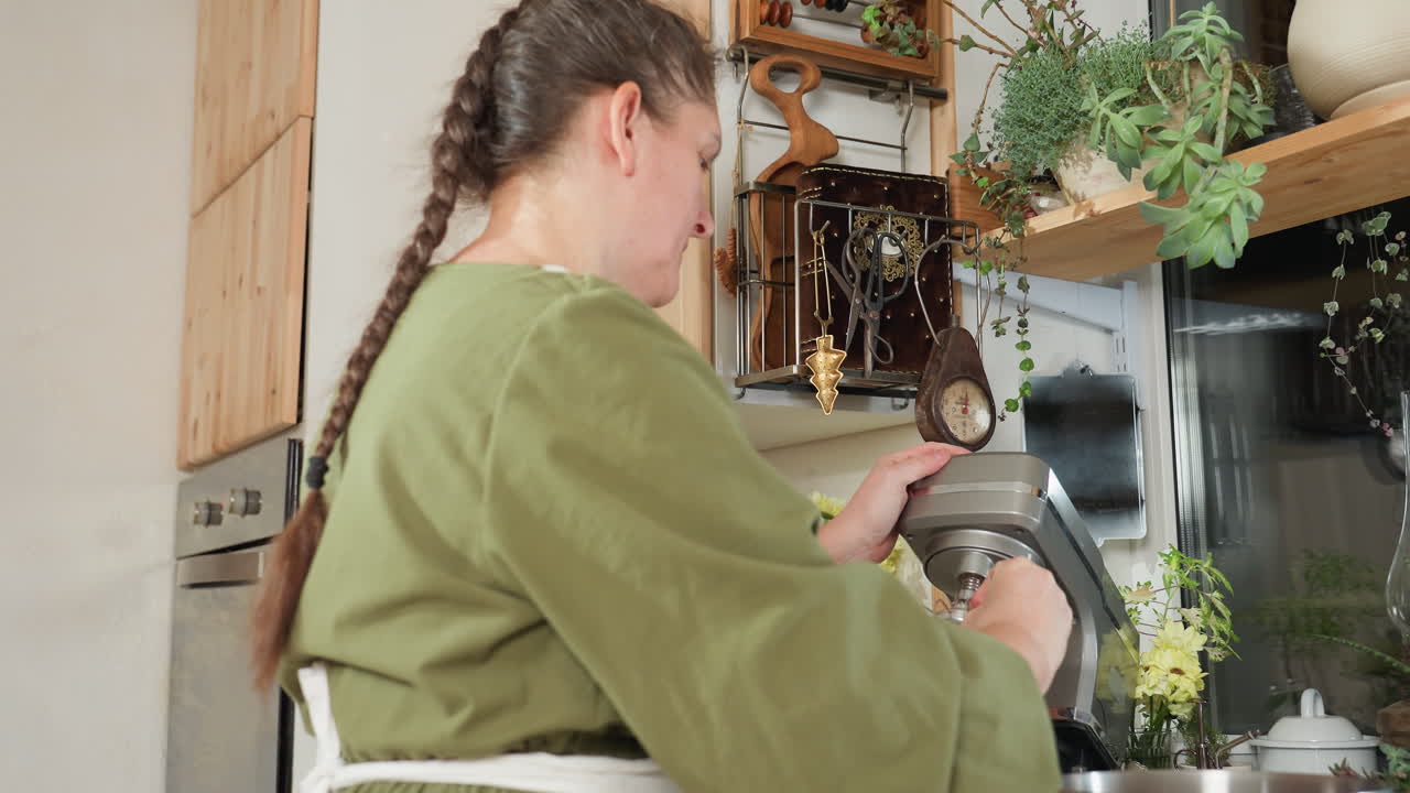 Woman in green dress and white apron attempts to fix whisk attachment into electric mixer inside bright vintage-style kitchen with wooden cabinets, indoor plants, utensils, and natural lighting