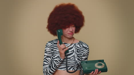 crazy girl with brown lush wig talking on wired vintage telephone of 80s, fooling making silly faces