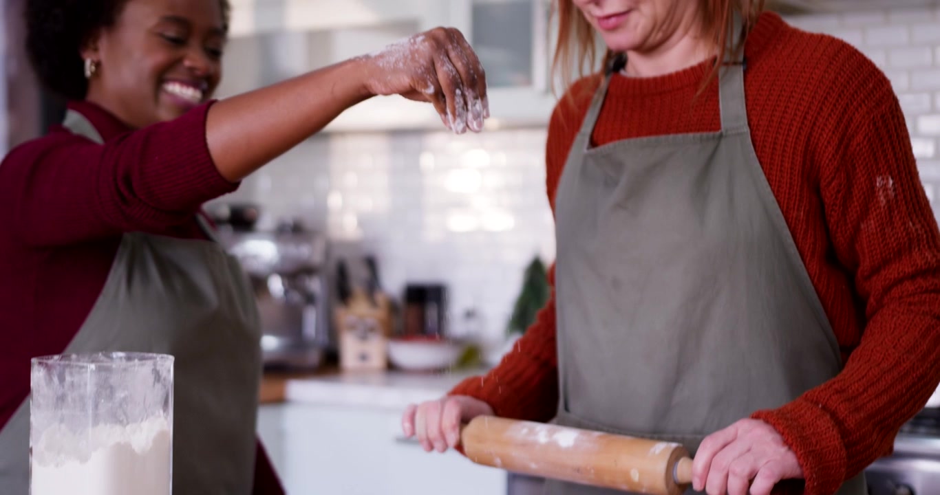 Diverse female friends lifting flour and using rolling pin on dough at home kitchen for baking