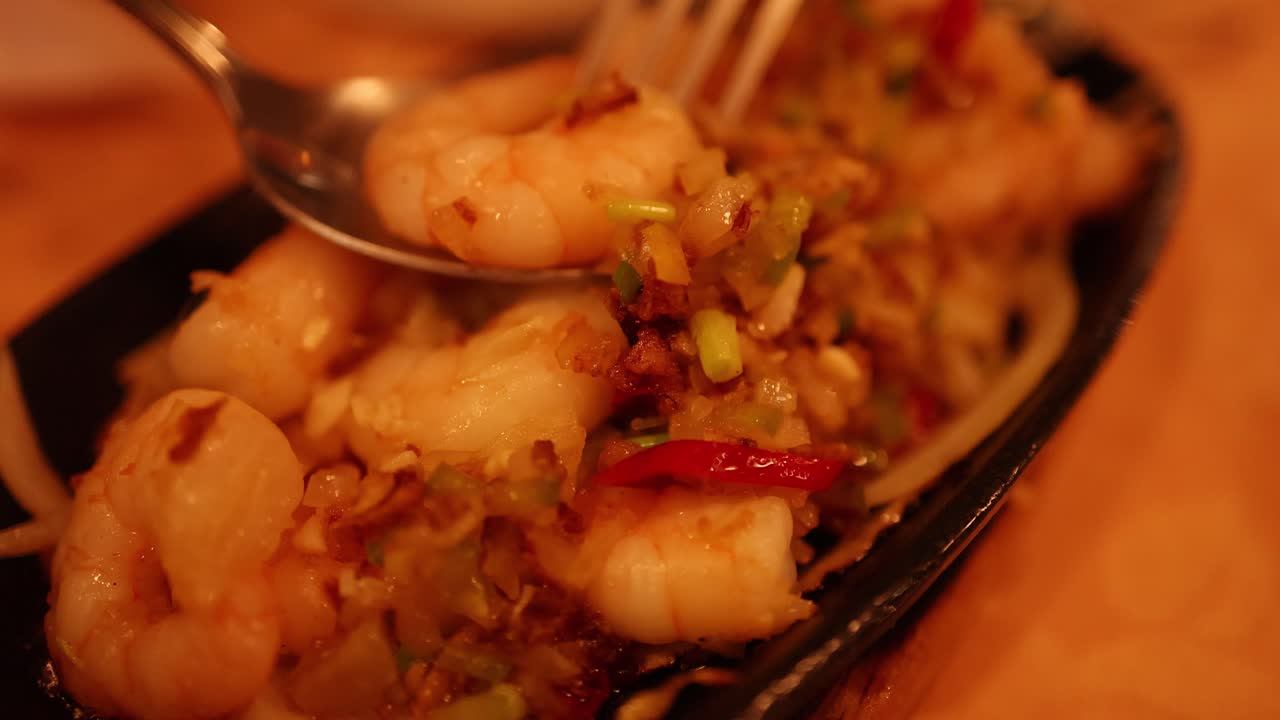 A fork lifts prawns and vegetables from a sizzling seafood platter under warm, ambient lighting in an Asian restaurant setting. Shallow depth of field