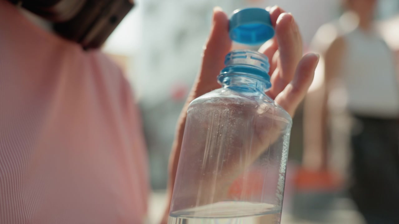 Woman holds clear plastic water bottle with blue cap unscrewing cap while standing outdoors in bright daylight. focus remains on her hand and transparent container showing liquid