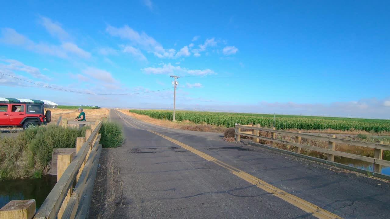 Pan of bridge over an irrigation canal; Visible are a corn field, alfalfa field, and a female with a camera walking toward her vehicle