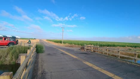 pan of bridge over an irrigation canal; visible are a corn field, alfalfa field, and a female with a camera walking toward her vehicle