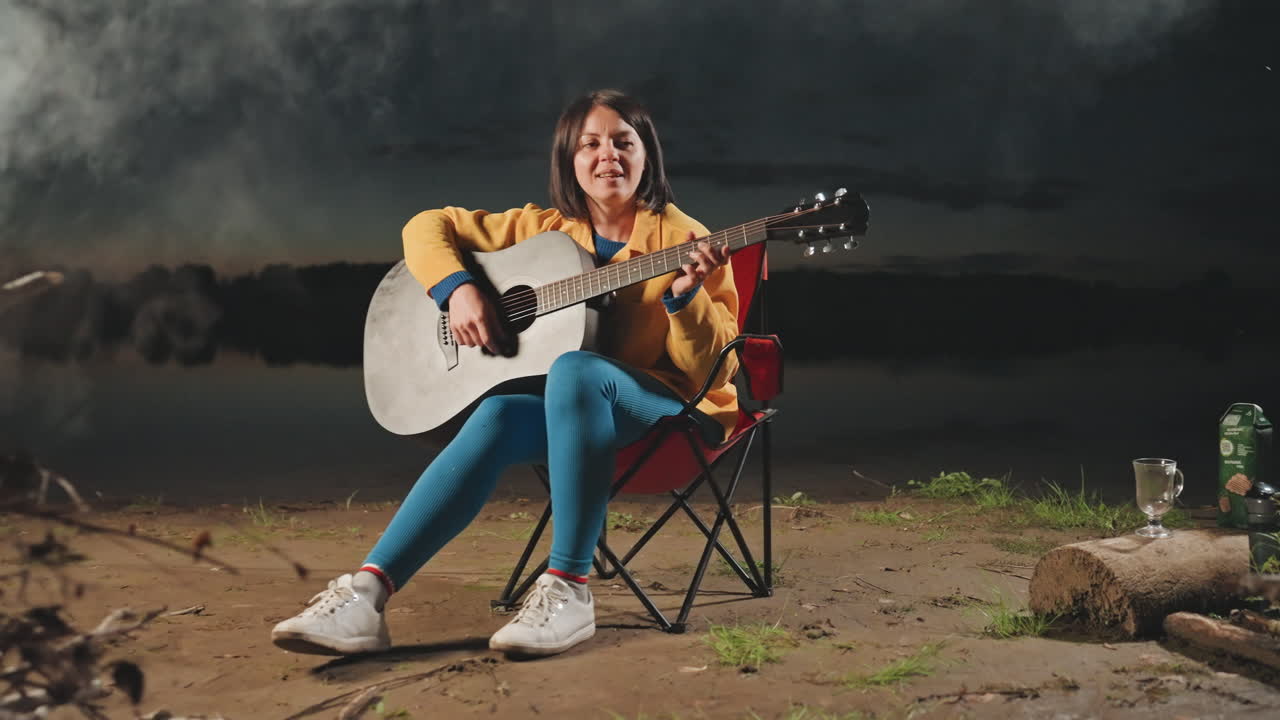 Totalbillede af hvid kvinde ved bål spiller akustisk guitar, bålrøg stiger op mod nattehimlen, brænde og flasker ved strandkanten, glad rejsende musikerstemning, frihed i åben natur.