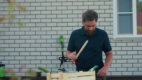 Bearded gardener by brick wall loads work tools into apron belt around waist, moving trowel with green handle from wooden crate on blue drum, organizing gear for yard tasks