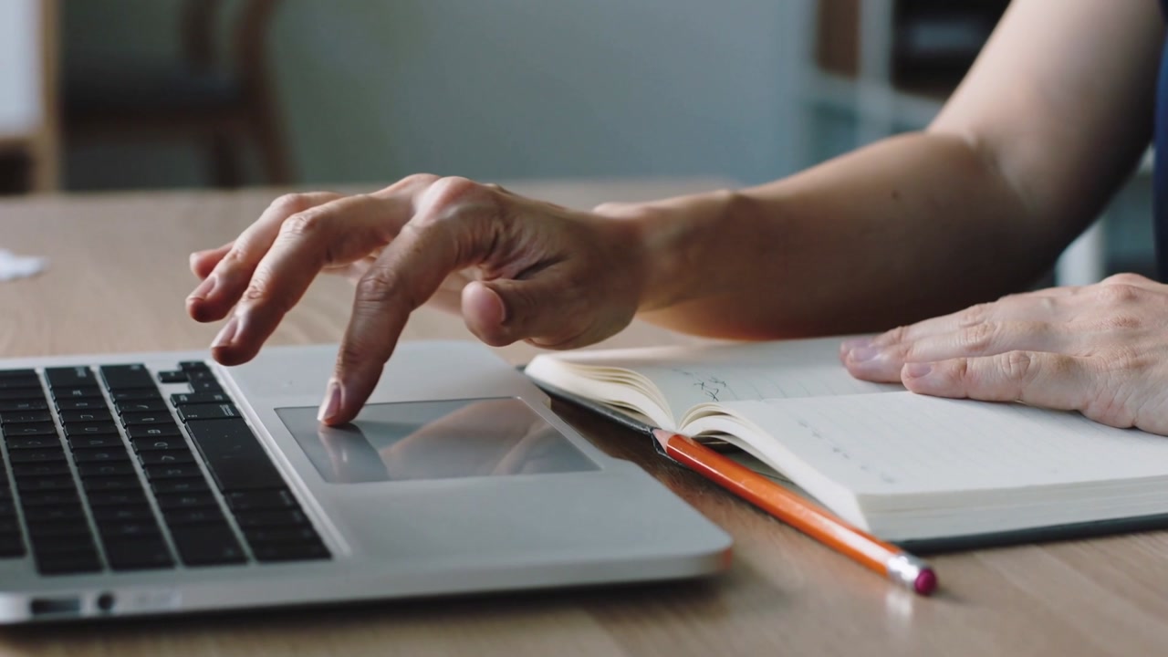 Hovering then touching woman in navy top using laptop trackpad at desk checking notes and pencil