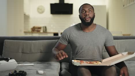 Afrikanischer-Mann-Sitzt-Auf-Dem-Sofa-In-Der-Offenen-Küche.-Schwarzer-Mann,-Der-Pizza-Isst.