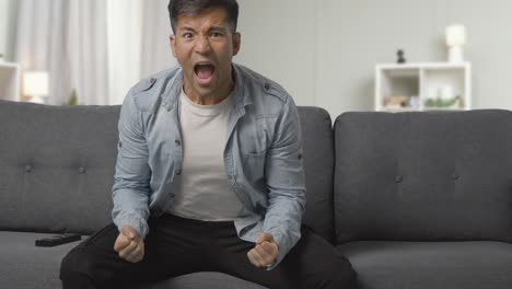 shot of excited young man sitting on sofa at home watching sport on tv and celebrating team goal or victory