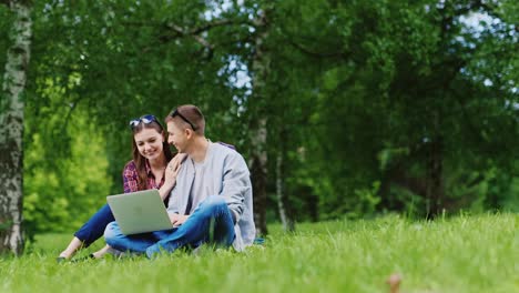 amigos de un hombre y una mujer sentados en el césped del parque disfrutan de un video hd de una laptop