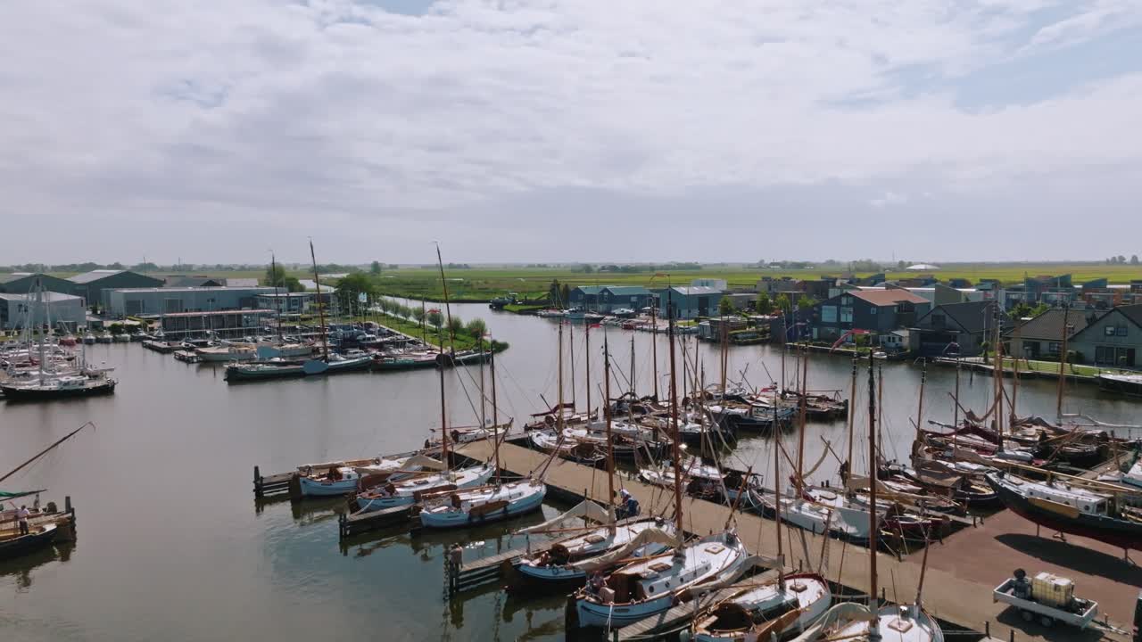 Drone zooms in while ascending, highlighting docked vintage sailboats and industrial waterfront at Heeg harbor