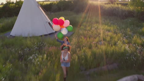 Drohnenaufnahme-Von-Zwei-Freundinnen,-Die-Auf-Einem-Musikfestival-Campen-Und-Mit-Luftballons-Durch-Das-Feld-Laufen