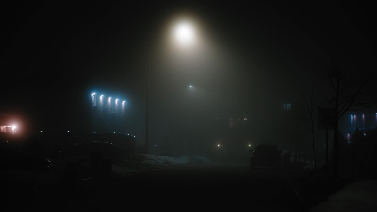 un hombre vestido con una chaqueta camina por una carretera vacía y oscura llena de niebla e iluminada por las luces de la calle