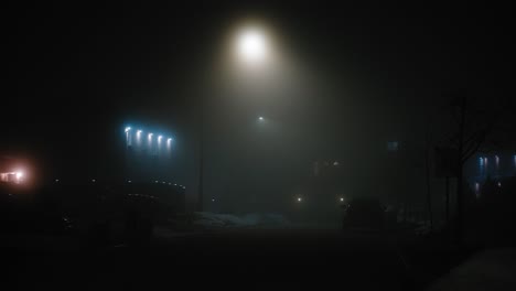 a man wearing a jacket walks down an empty and dark road filled with fog and illuminated by street lights
