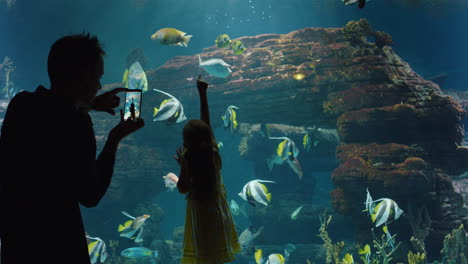 family at aquarium father taking photo of daughter using smartphone little girl  looking at fish swimming in corel reef with dad sharing fun at oceanarium on social media