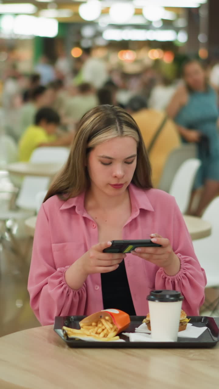 dama blanca en vestido rosa de pie en una mesa de madera en un restaurante ocupado tomando una foto de su comida, se sienta para tomar una foto de sus comidas, con el fondo ligeramente borroso con la gente
